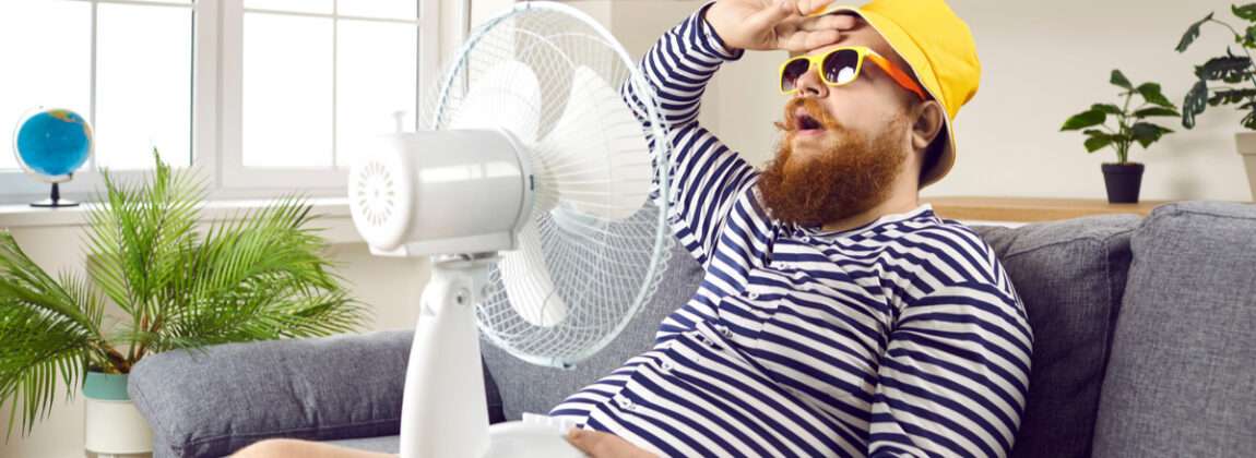 扇風機を手にソファに座り、英語で話すひげを生やした男性。