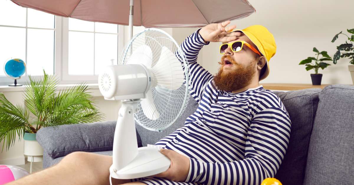 扇風機を手にソファに座り、英語で話すひげを生やした男性。