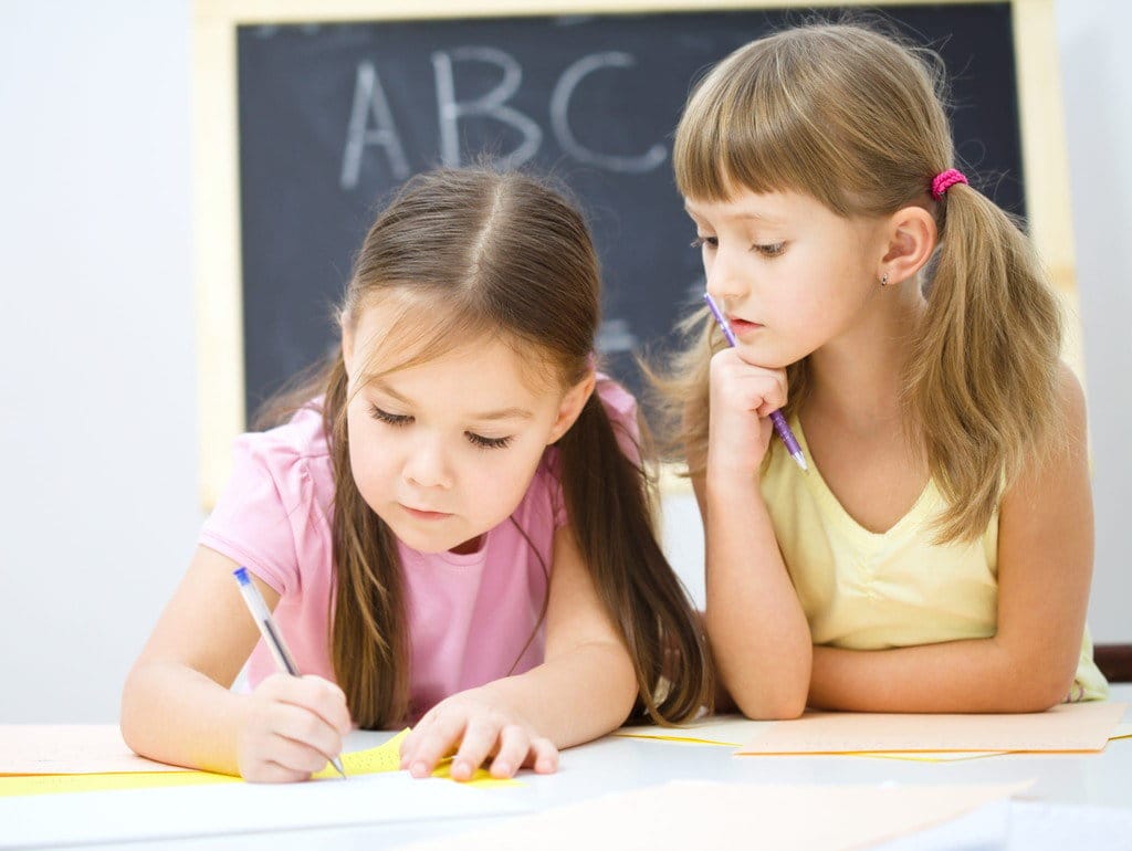 二人の女の子が机に向かって日本語（英語）の会話を練習したり、紙に書いたりしています。