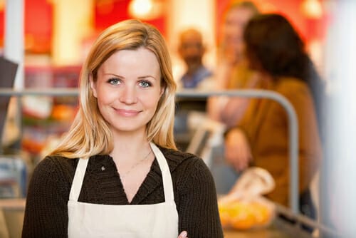 エプロンを着て食料品店に立ち、顧客と英会話を交わし、英語で対応する女性。