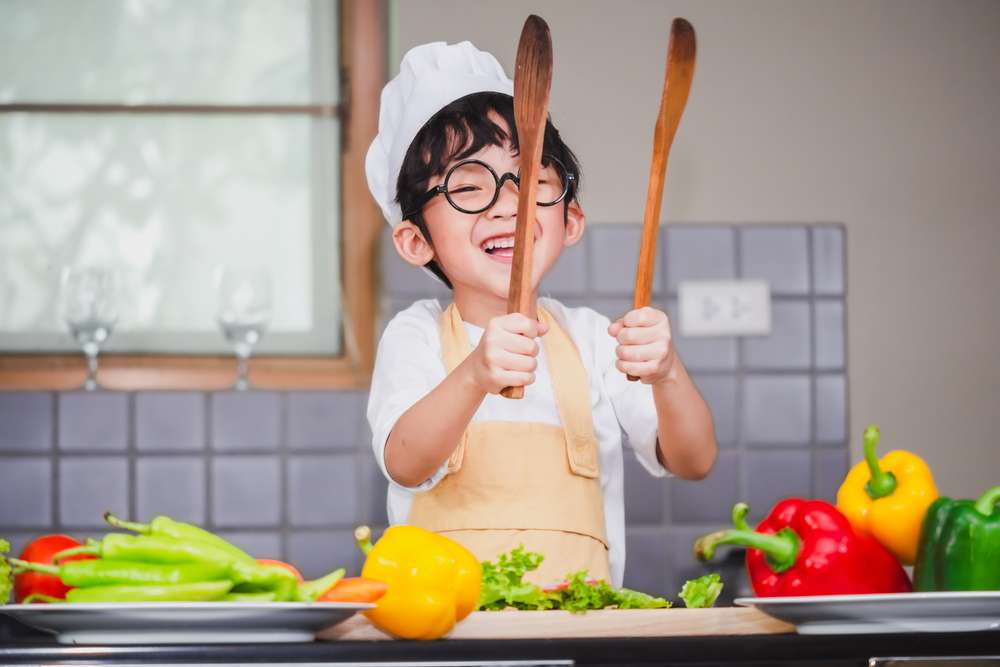 キッチンで 2 つの木のスプーンを持ち、料理を学ぶ少年。