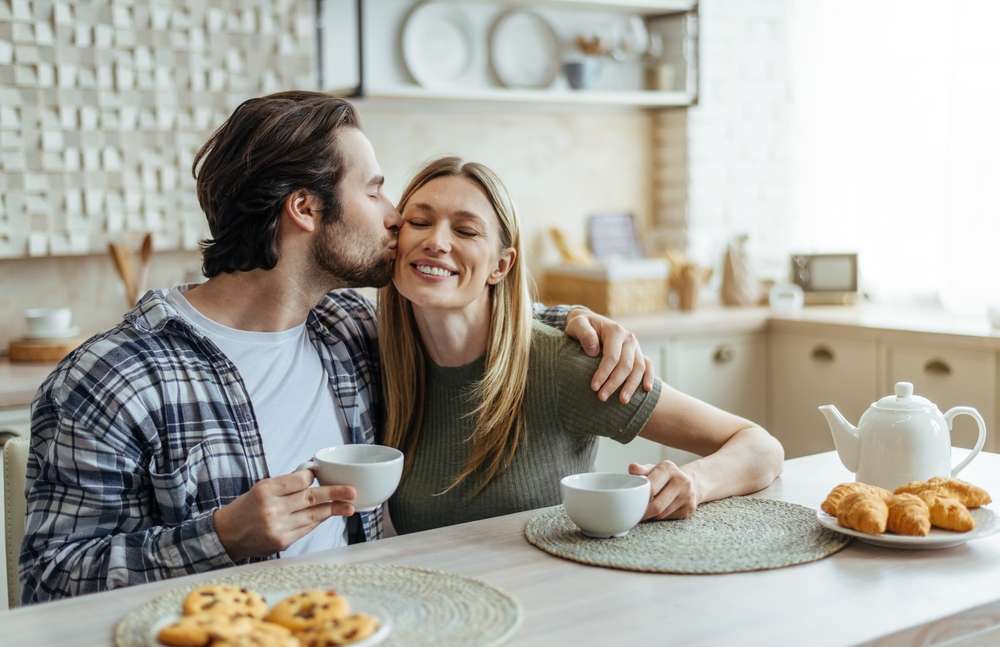 男性が女性の頬にキスをしながら、キッチンのテーブルで紅茶とペストリーを楽しんでいます。2人とも温かく微笑みながら英語で会話しています。