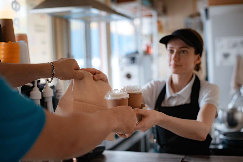 帽子をかぶったバリスタが、カフェのカウンターで英語を話す客にバッグとコーヒーカップ2つを渡している。