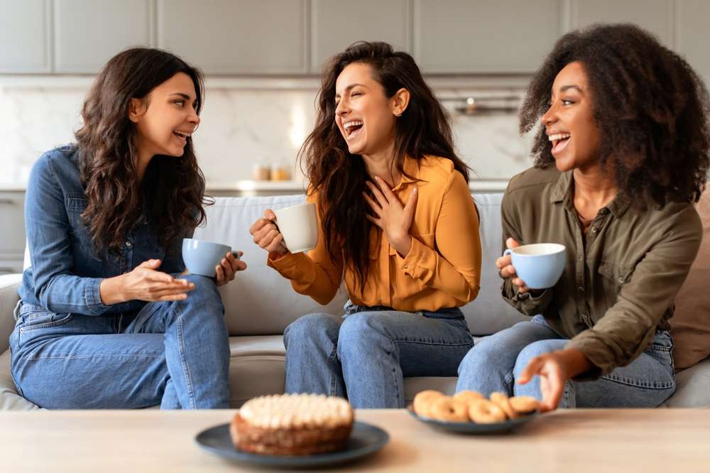 3人の女性がキッチンのソファーに座ってコーヒーを飲みながら笑っています。目の前のテーブルにはケーキとクッキーが置いてあります。彼女たちは甘いお菓子を味わいながら英語で会話をしています。