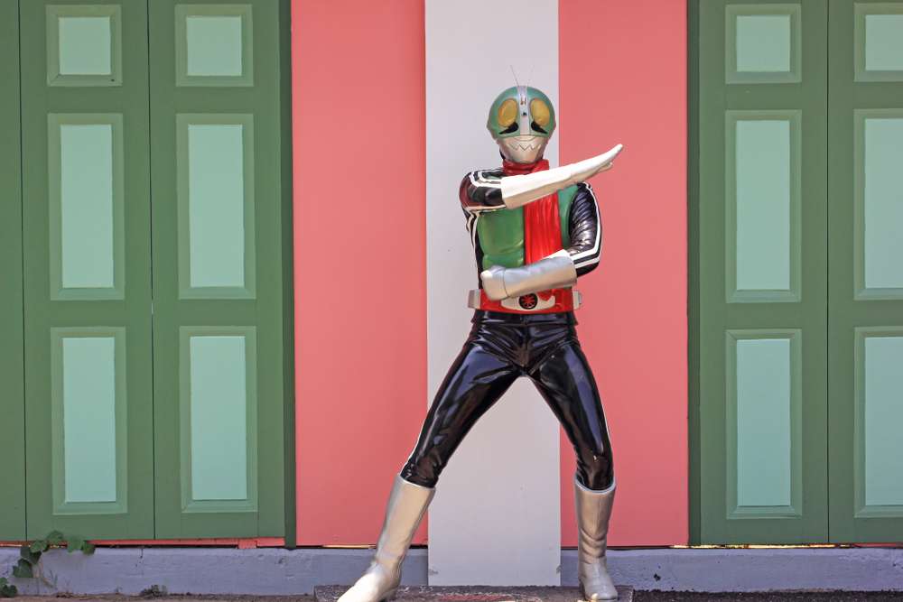 緑、黒、赤のスーパーヒーローのコスチュームを着た人が、緑のドアが付いたカラフルな壁の前で腕を組んでポーズをとり、英語で書かれたエンブレムを誇らしげに見せています。