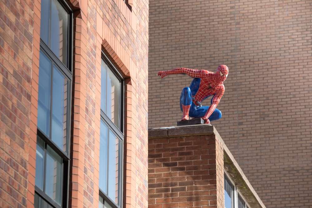 スパイダーマンに扮した人物がレンガ造りの建物の端にしゃがみ込んでおり、背景には別の建物が見え、今にも動き出しそうな様子だ。