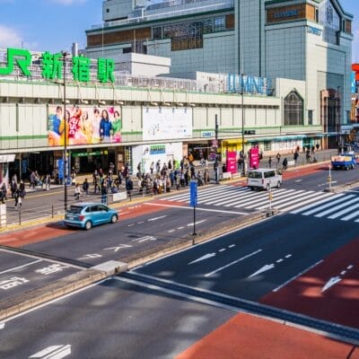 東京の JR 新宿駅前にある繁華街。晴れた日には、高層ビルに囲まれ歩行者と車両が行き交う。活気に満ちた環境は、地元のカフェでの英会話マンツーマンセッションに最適な背景を提供します。.