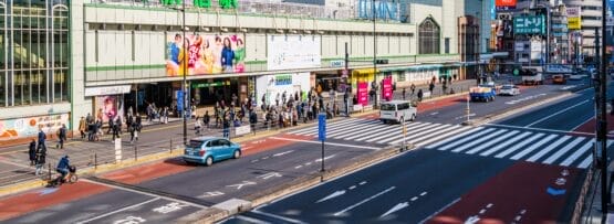 東京の JR 新宿駅前にある繁華街。晴れた日には、高層ビルに囲まれ歩行者と車両が行き交う。活気に満ちた環境は、地元のカフェでの英会話マンツーマンセッションに最適な背景を提供します。.