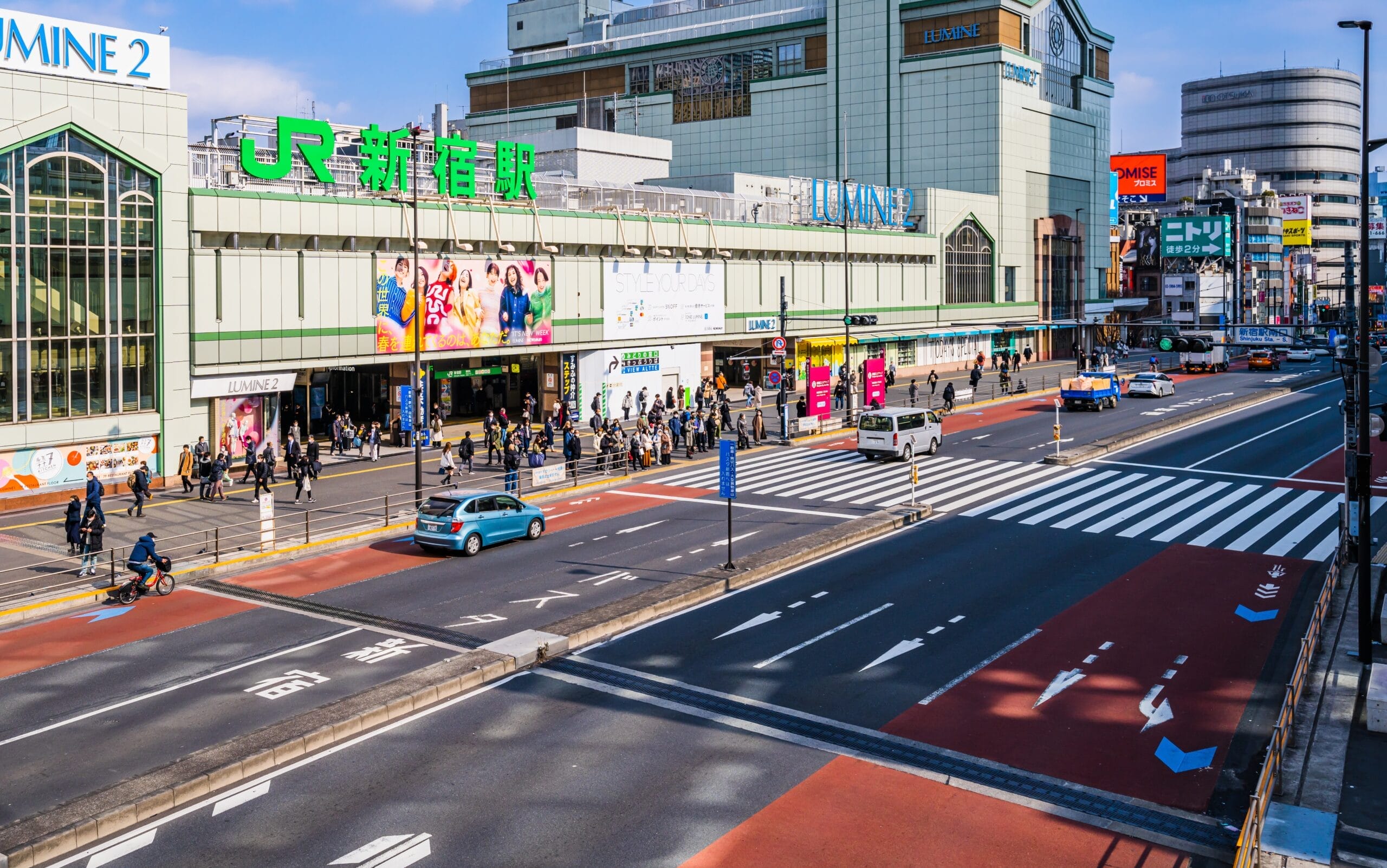 東京の JR 新宿駅近くのにぎやかな街並み。歩行者が交差点を渡り、車両が近代的な店舗の横を移動する。この都市のエネルギーの中で、英会話マンツーマンの機会が繁栄し、活気に満ちた都市景観に溶け込みます。.