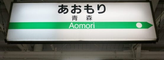 駅の標識には、ローマ字と日本語で「Aomori」と書かれており、方向を示す緑の線と白い円で「Aomori・弘前」と表示されています。すべて明るい屋内環境に設置されています。.