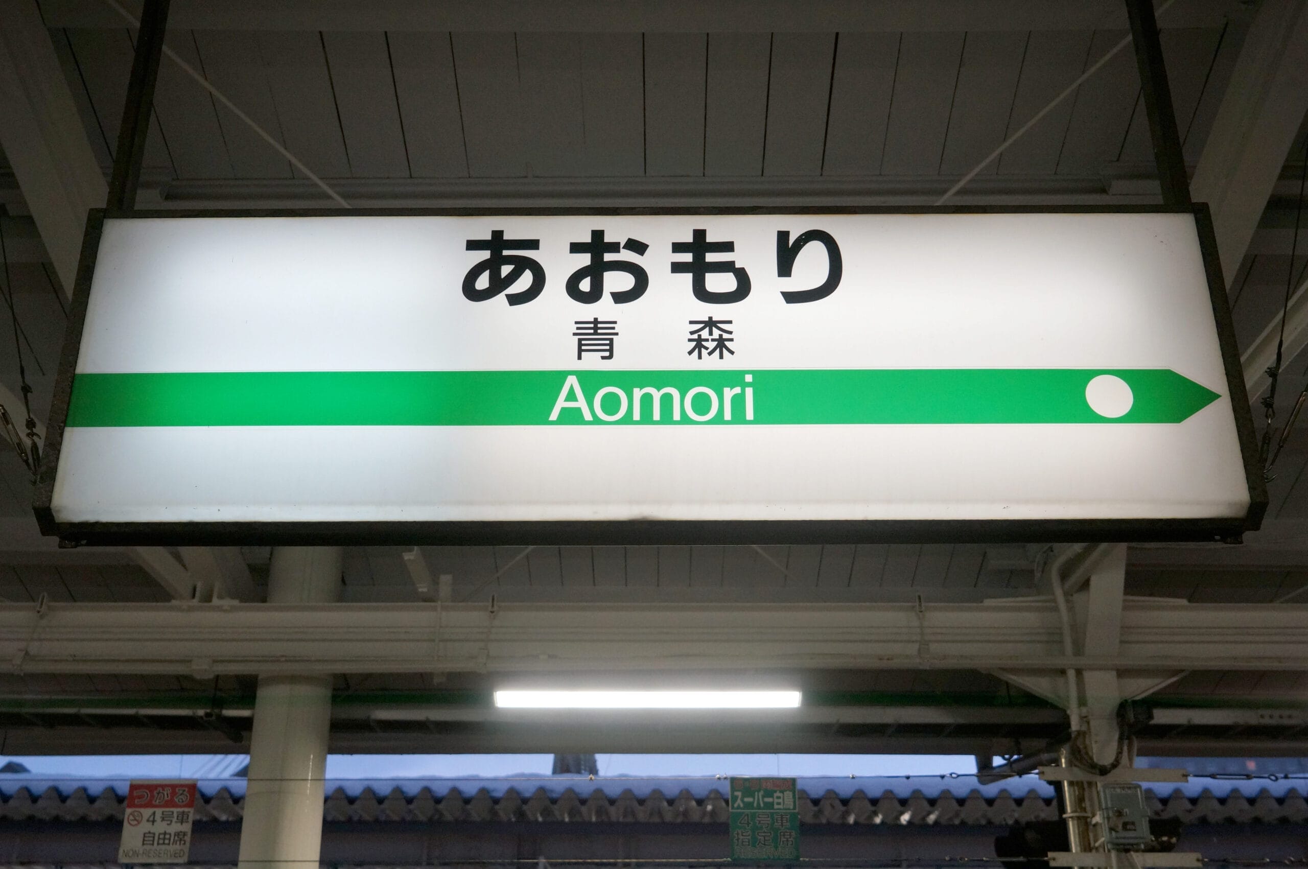駅の標識には、ローマ字と日本語で「Aomori」と書かれており、方向を示す緑の線と白い円で「Aomori・弘前」と表示されています。すべて明るい屋内環境に設置されています。.
