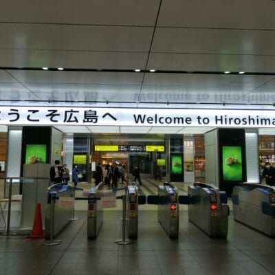 駅の改札口には、日本語と英語で「広島へようこそ」と書かれた看板が掲げられ、訪れる人々を温かく迎えている。背景には乗客の賑わいが見られ、広島の活気ある雰囲気が伝わってくる。.