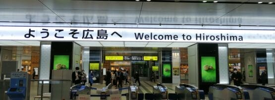 駅の改札口には、日本語と英語で「広島へようこそ」と書かれた看板が掲げられ、訪れる人々を温かく迎えている。背景には乗客の賑わいが見られ、広島の活気ある雰囲気が伝わってくる。.