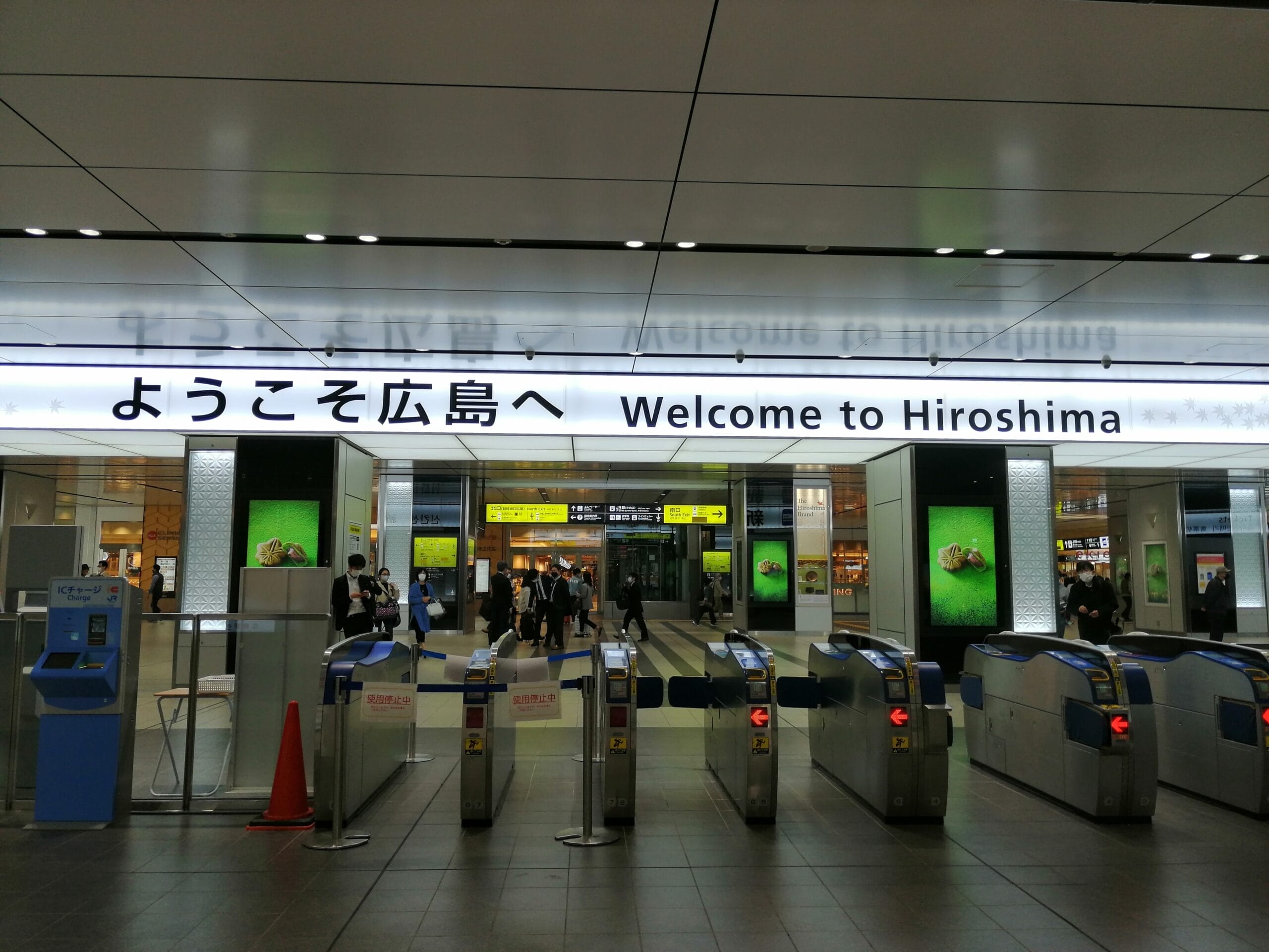 駅の改札口には、日本語と英語で「広島へようこそ」と書かれた看板が掲げられ、訪れる人々を温かく迎えている。背景には乗客の賑わいが見られ、広島の活気ある雰囲気が伝わってくる。.