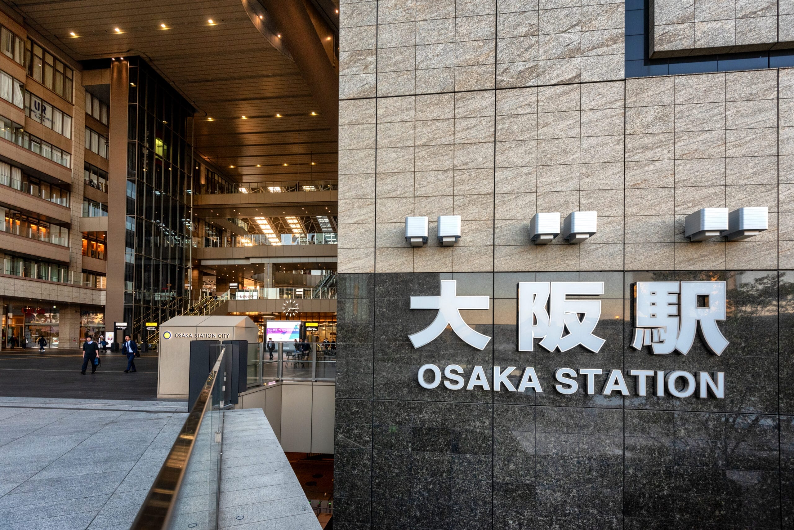 大阪駅の外観では、石垣に大きな看板が掲げられ、人々が近くを散策しています。街の活気を感じさせる近代的な建物が背景にそびえ立っています。近くには、大阪の伝統と現代的なコミュニケーションの融合を反映した英会話マンマンツーセンターがあります。.