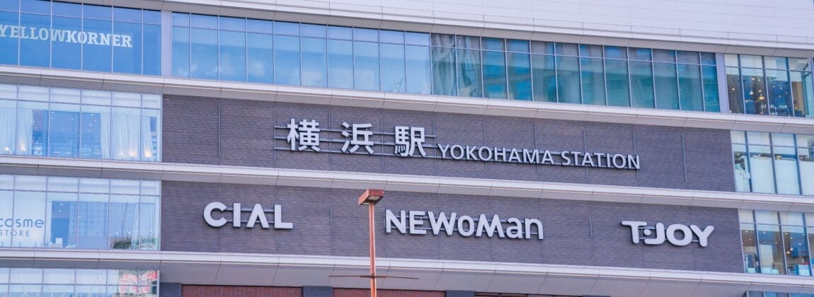 この画像は日本の横浜駅の外観を示しており、高層ビルのファサードに対してCIAL、NEWoMan、TJOYの看板が強調されています。活気あふれる横浜の街にあるこの賑やかなエリアは、文化的な楽しみを探索したり、近くで英会話マンツーマンのレッスンを見つけたりするのに理想的な場所です。.