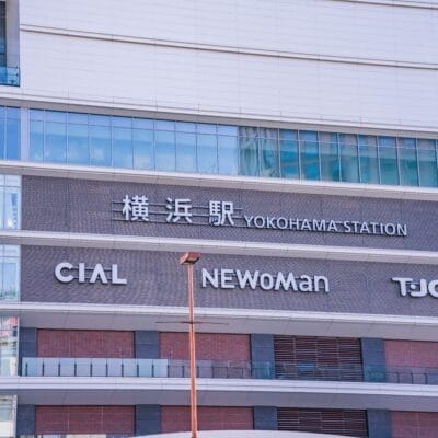 この画像は日本の横浜駅の外観を示しており、高層ビルのファサードに対してCIAL、NEWoMan、TJOYの看板が強調されています。活気あふれる横浜の街にあるこの賑やかなエリアは、文化的な楽しみを探索したり、近くで英会話マンツーマンのレッスンを見つけたりするのに理想的な場所です。.