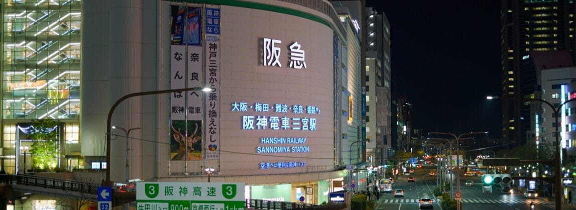 夜の賑やかな街路には、阪急電鉄の電飾看板や日本語の道路標識が灯り、街を明るく照らしています。高層ビルの間を車が行き交い、神戸三宮の看板が目立つように立ち、活気に満ちた都会の風景の中を旅人を案内しています。.