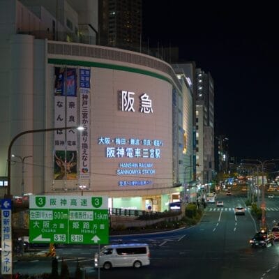 夜の賑やかな街路には、阪急電鉄の電飾看板や日本語の道路標識が灯り、街を明るく照らしています。高層ビルの間を車が行き交い、神戸三宮の看板が目立つように立ち、活気に満ちた都会の風景の中を旅人を案内しています。.