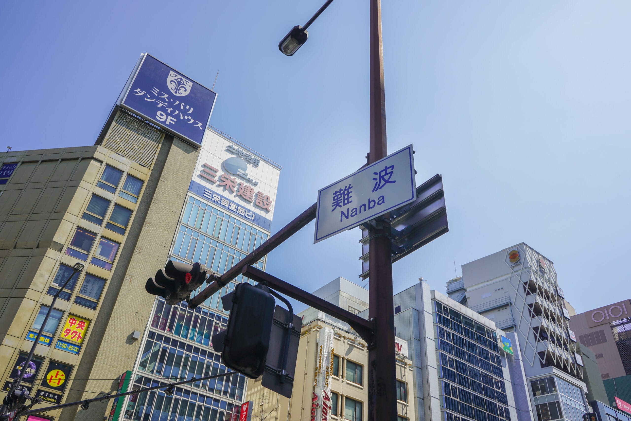 難波・なんばのストリートビュー。澄んだ青空を背景に、英会話マンツーマンレッスンを宣伝する看板と信号機のある高層ビルが特徴。.
