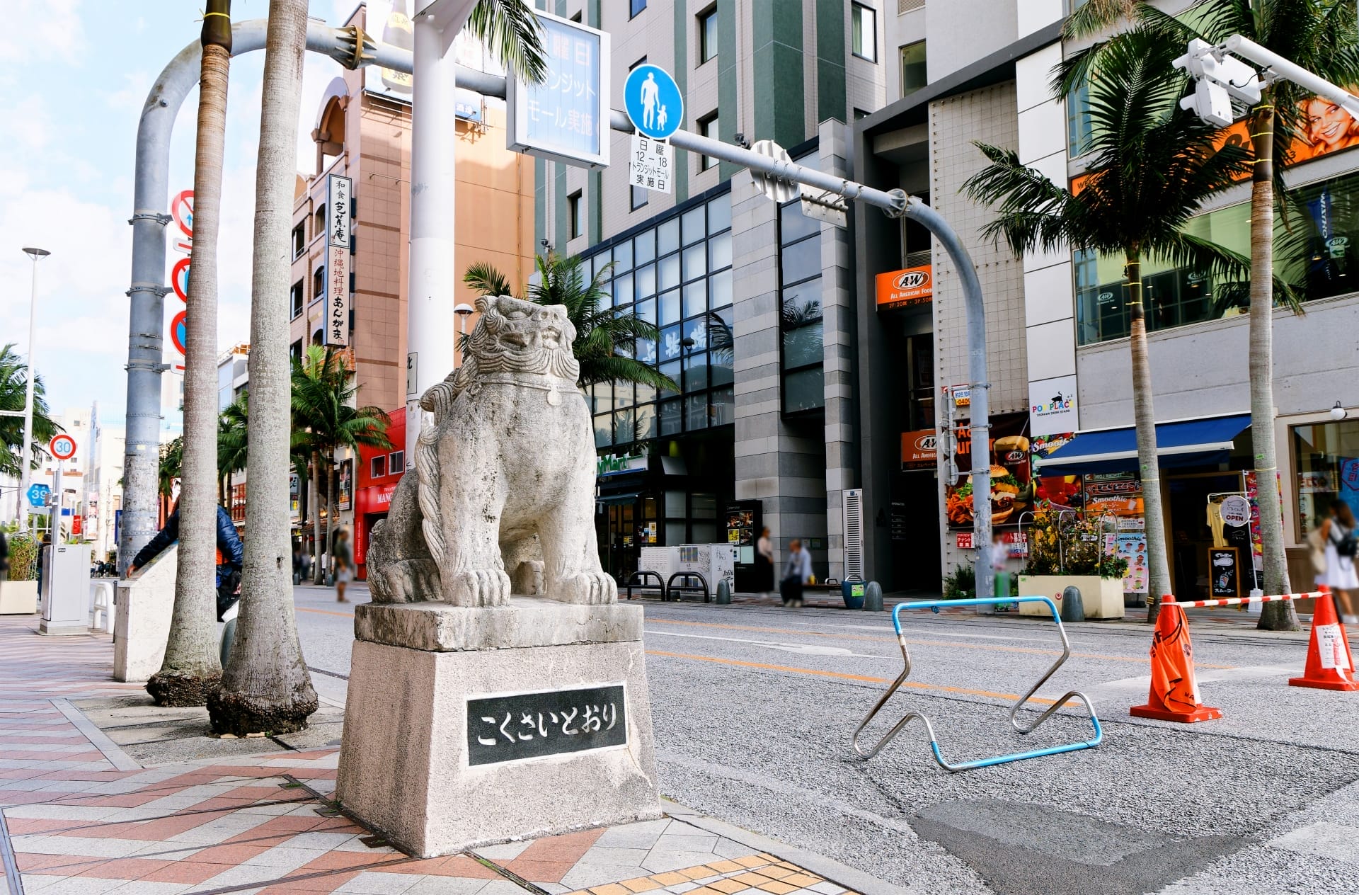 揺れるヤシの木、近代的な建物、活気に満ちた交通標識に囲まれた沖縄・那覇の賑やかな一角に、ライオンの石像が堂々と立っています。近くでは通勤する地元の人々や英会話スクールマンツーマンのセッションに向かう観光客が静かに目撃されています。.
