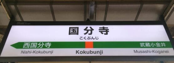 駅名標には「国分寺」が大きく表示され、その下に「西国分寺」と「武蔵小金井」が都会的な背景に囲まれています。近くには、国分寺の中心部にある英会話スクール マンツーマンが個人に合わせた英語レッスンを提供しています。.