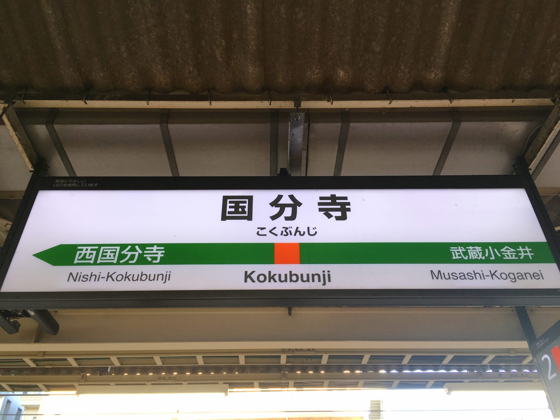 駅名標には「国分寺」が大きく表示され、その下に「西国分寺」と「武蔵小金井」が都会的な背景に囲まれています。近くには、国分寺の中心部にある英会話スクール マンツーマンが個人に合わせた英語レッスンを提供しています。.