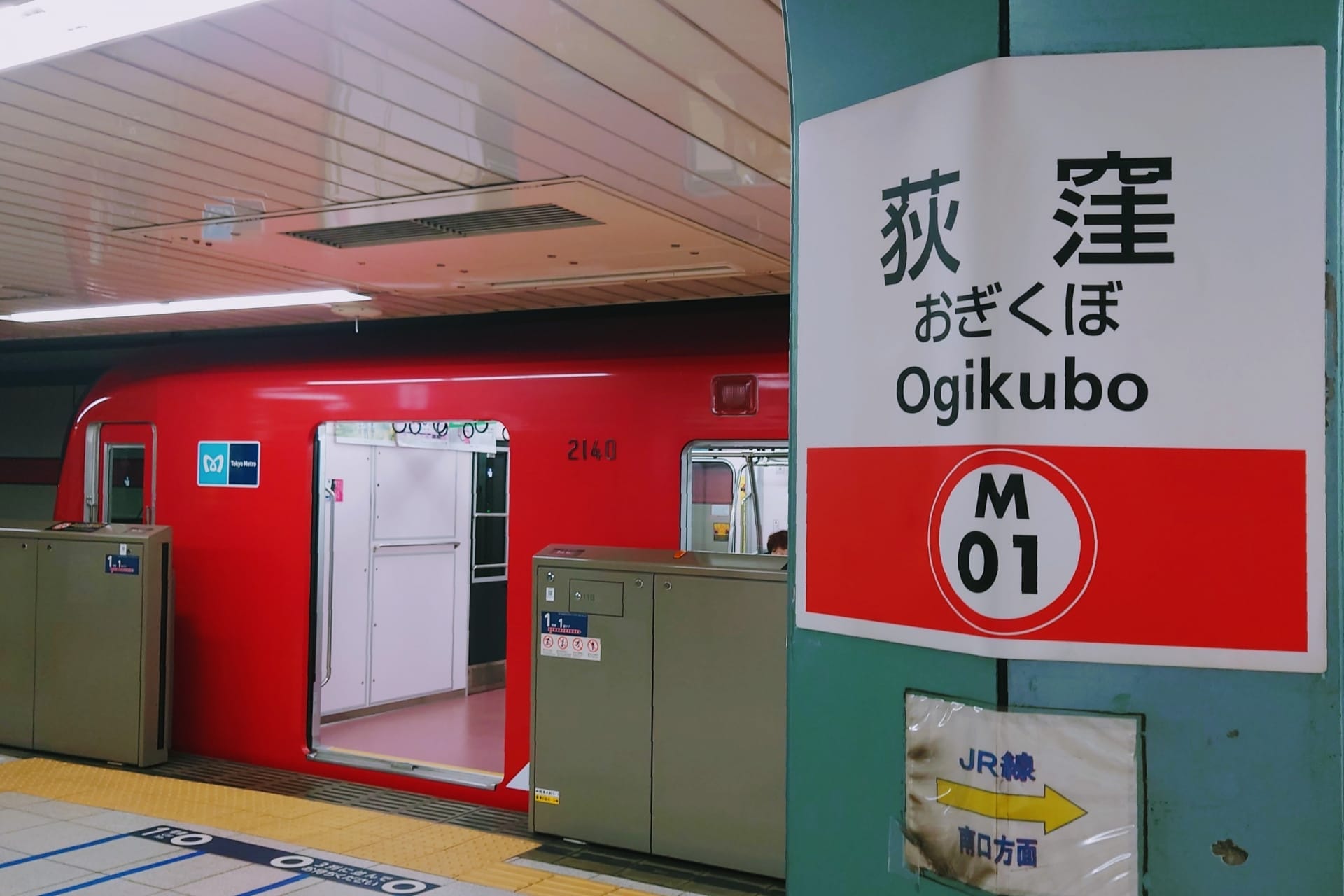 荻窪駅に赤い電車が止まっています。近くの標識には「荻窪」と駅コードM01が英語と日本語で表示されています。このプラットフォームに隣接する英会話スクールマンツーマンは、言語スキルを求める通勤者に個別のレッスンを提供しています。.