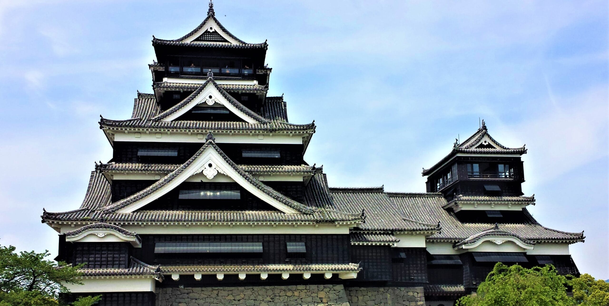 多層の屋根と木製の要素を備えた伝統的な日本の城。熊本の青い空を背景に、前景には緑が広がっています。.