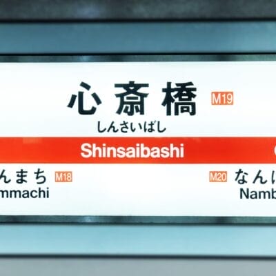 心斎橋の地下鉄駅名標には日本語と英語で「心斎橋」と表示されています。英会話スクールマンツーマンの近くに位置し、駅コードM19、M18、M20とともに「本町」「難波」への道順を掲載しています。.