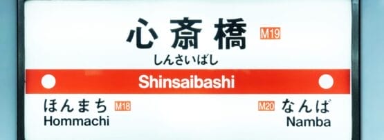 心斎橋の地下鉄駅名標には日本語と英語で「心斎橋」と表示されています。英会話スクールマンツーマンの近くに位置し、駅コードM19、M18、M20とともに「本町」「難波」への道順を掲載しています。.