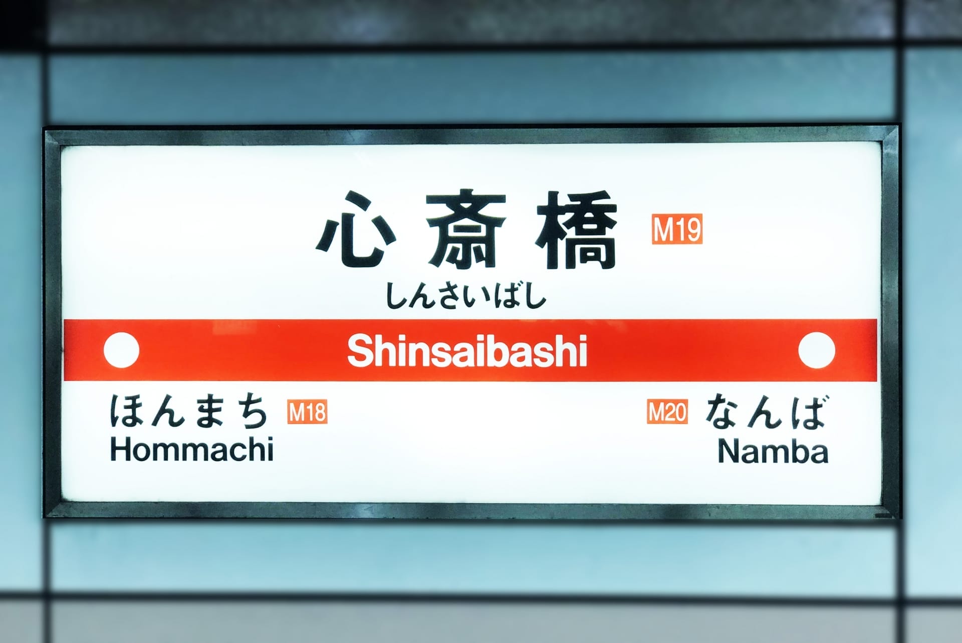 心斎橋の地下鉄駅名標には日本語と英語で「心斎橋」と表示されています。英会話スクールマンツーマンの近くに位置し、駅コードM19、M18、M20とともに「本町」「難波」への道順を掲載しています。.
