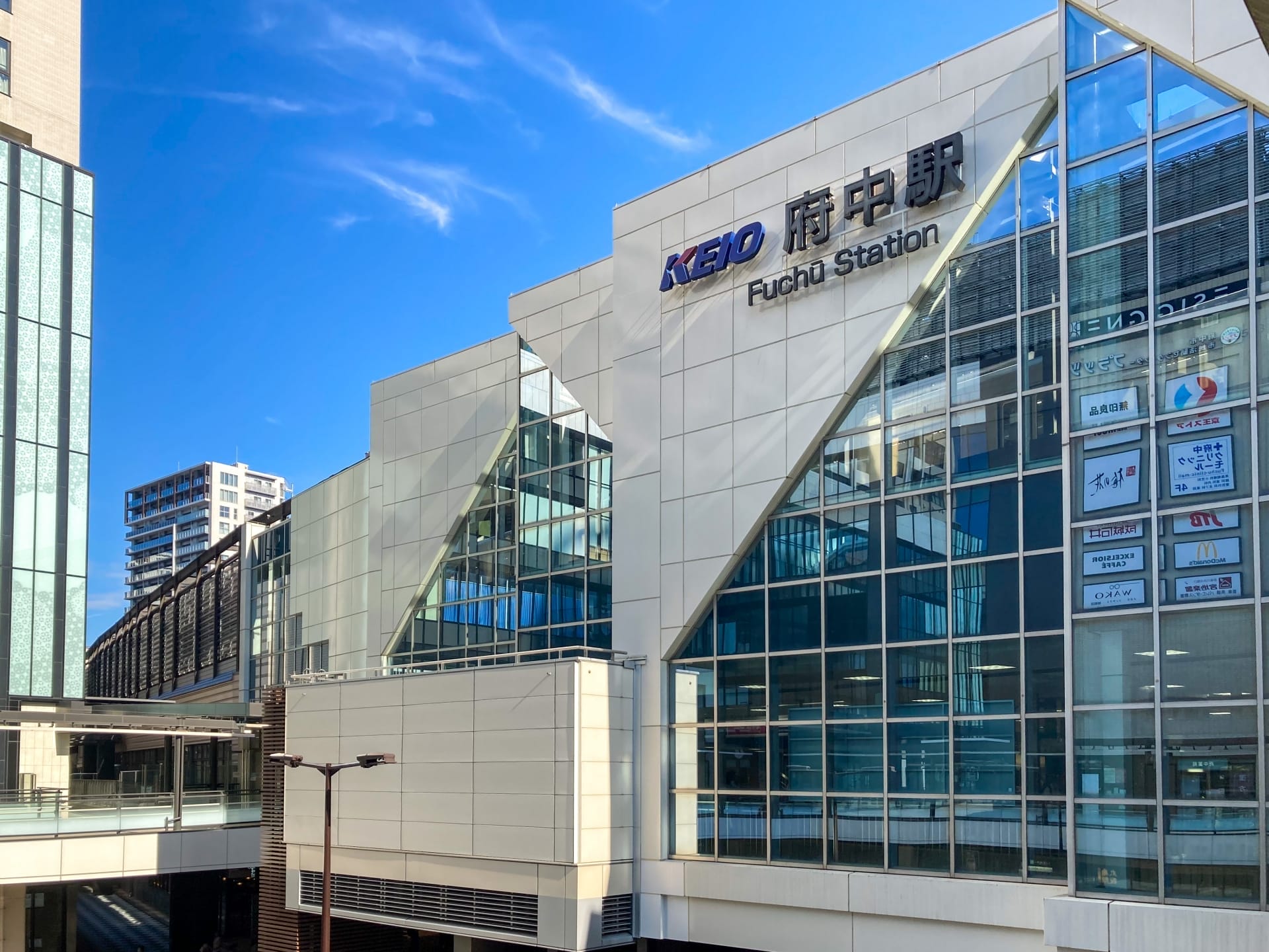 京王府中駅の外観。澄み切った青空を背景に、大きな窓と企業名が書かれた看板のあるモダンな建築が特徴。.