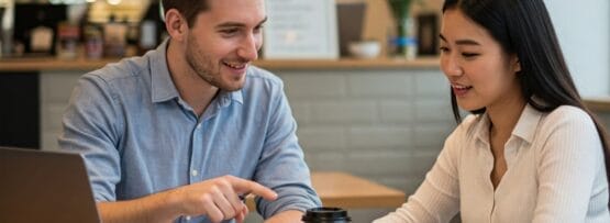 男性と女性がカフェのテーブルにノートパソコン、コーヒー、書類を持って座り、話し合いをしながら互いに微笑んでいます。.